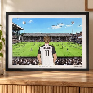 May include: A framed illustration of a soccer stadium featuring a person from behind wearing a white and black jersey with the name CHARLIE and the number 11 printed on the back. The text Our family. Our club. is printed at the bottom of the print.
