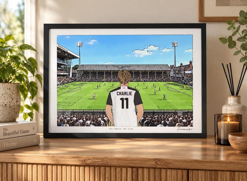 May include: A framed illustration of a soccer stadium featuring a person from behind wearing a white and black jersey with the name CHARLIE and the number 11 printed on the back. The text Our family. Our club. is printed at the bottom of the print.