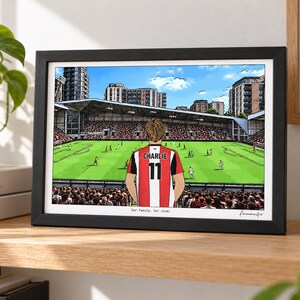 May include: A framed horizontal print featuring a cartoon illustration of a football player wearing a red and white striped jersey with the name CHARLIE and number 11 on the back, standing before a stadium pitch with the text Our family. Our club.
