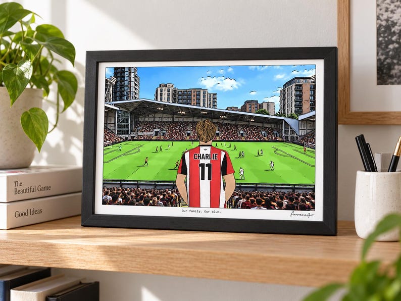 May include: A framed horizontal print featuring a cartoon illustration of a football player wearing a red and white striped jersey with the name CHARLIE and number 11 on the back, standing before a stadium pitch with the text Our family. Our club.