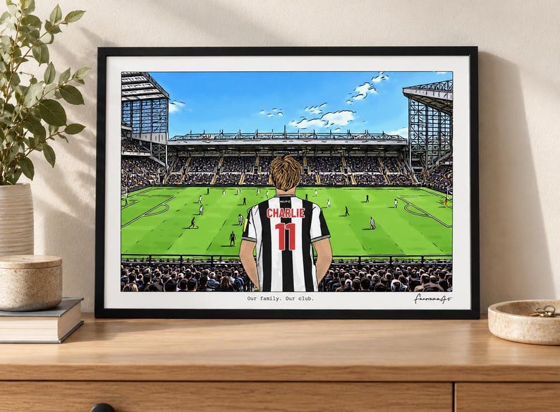 May include: A framed art print depicting a soccer stadium view from the stands. A person stands in the foreground wearing a black and white striped jersey with the name CHARLIE and the number 11 on the back. Text reads Our family. Our club.