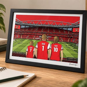 AI-created lifestyle mockup of a black framed A4 Arsenal family print standing on a wooden desk beside a notebook and mug, featuring Jamie 7, Dad 1 and Mum 10 with the red-sky Pitch Side background.