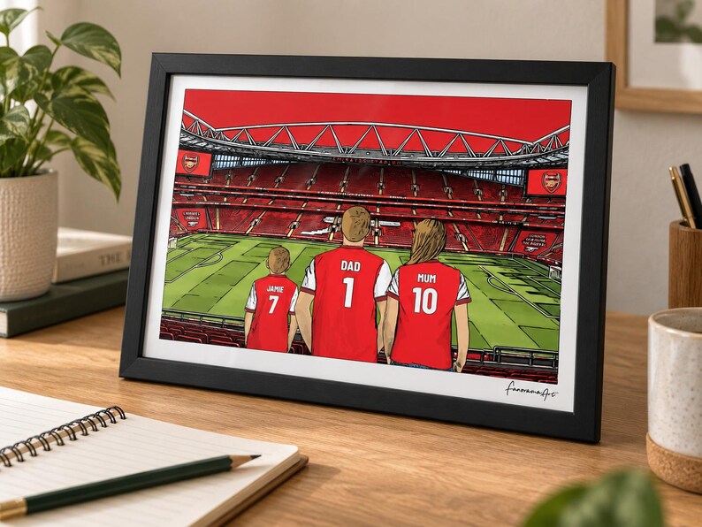 AI-created lifestyle mockup of a black framed A4 Arsenal family print standing on a wooden desk beside a notebook and mug, featuring Jamie 7, Dad 1 and Mum 10 with the red-sky Pitch Side background.