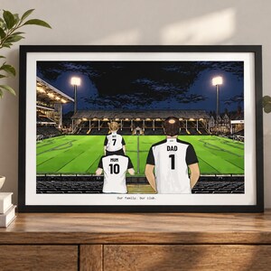 May include: A framed illustration of a family at a soccer stadium at night. The figures wear white and black jerseys labeled Mum 10, Dad 1, and Ivy 7. The stadium features bright floodlights, a green pitch, and dark stands. Text reads Our family. Our club.