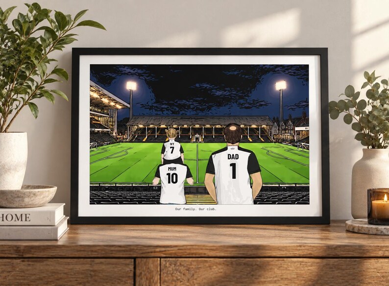 May include: A framed illustration of a family at a soccer stadium at night. The figures wear white and black jerseys labeled Mum 10, Dad 1, and Ivy 7. The stadium features bright floodlights, a green pitch, and dark stands. Text reads Our family. Our club.