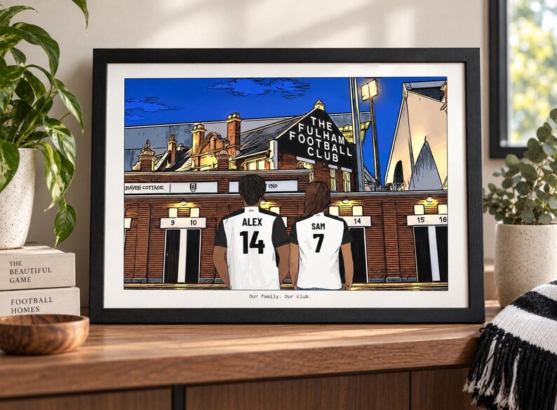 May include: A framed illustration of two people wearing white and black soccer jerseys with the names ALEX and SAM and numbers 14 and 7 on the back, standing before The Fulham Football Club stadium at night with the text Our family. Our club.