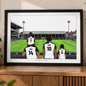 May include: A framed illustration of a family of four standing in a soccer stadium. The figures wear white and black jerseys with the names Max, Dad, Mum, and Ava and numbers 7, 14, 10, and 9. The text Our family. Our club. is printed at the bottom.
