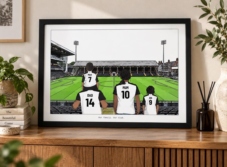 May include: A framed illustration of a family of four standing in a soccer stadium. The figures wear white and black jerseys with the names Max, Dad, Mum, and Ava and numbers 7, 14, 10, and 9. The text Our family. Our club. is printed at the bottom.