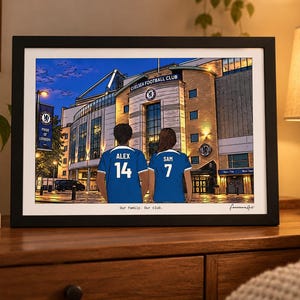 Puede incluir: Una ilustraci&oacute;n enmarcada de dos personas con camisetas azules del Chelsea Football Club con los nombres ALEX y SAM y los n&uacute;meros 14 y 7 en la espalda, frente al estadio. El texto Our family. Our club. est&aacute; impreso en la parte inferior.