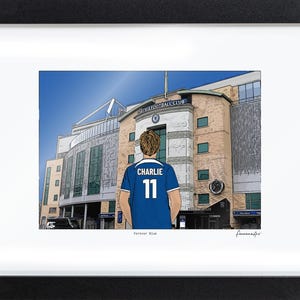 Puede incluir: Una ilustraci&oacute;n enmarcada de una persona con una camiseta de f&uacute;tbol azul con el nombre CHARLIE y el n&uacute;mero 11 en la espalda, frente a un gran estadio con el texto CHELSEA FOOTBALL CLUB y las palabras Forever Blue debajo.