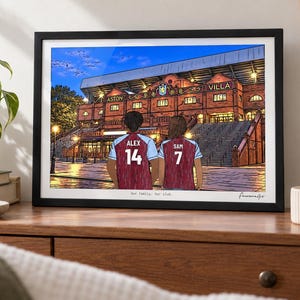 May include: A framed illustration of two people wearing claret and blue soccer jerseys with the names ALEX 14 and SAM 7, standing before the Aston Villa stadium at dusk. The text Our family. Our club. is printed at the bottom of the artwork.
