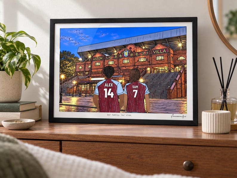 May include: A framed illustration of two people wearing claret and blue soccer jerseys with the names ALEX 14 and SAM 7, standing before the Aston Villa stadium at dusk. The text Our family. Our club. is printed at the bottom of the artwork.