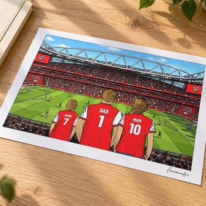 AI-created lifestyle mockup of an unframed A4 Arsenal family print lying on an oak desk beside a cream notebook and coffee, featuring Jamie 7, Dad 1 and Mum 10 overlooking a match in the In game background.
