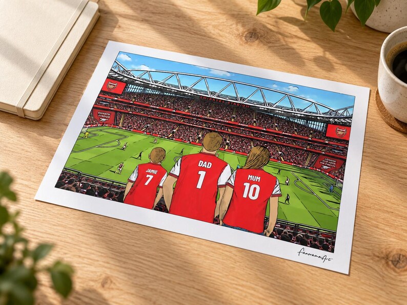 AI-created lifestyle mockup of an unframed A4 Arsenal family print lying on an oak desk beside a cream notebook and coffee, featuring Jamie 7, Dad 1 and Mum 10 overlooking a match in the In game background.