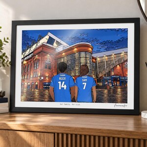 May include: A framed digital illustration depicting two people from behind wearing blue soccer jerseys with the names ALEX and SAM and numbers 14 and 7, standing before a large brick stadium. The text Our family. Our club. is printed below.