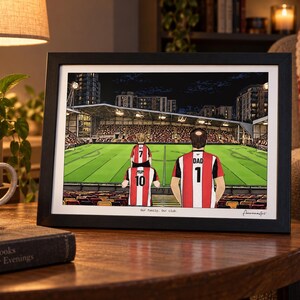 May include: A framed illustration of a family at a football stadium. The artwork depicts three people wearing red and white striped jerseys with the text MUM 10, DAD 1, and a child with 7. The text Our family: Our club is printed at the bottom.