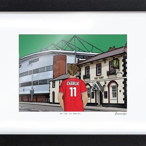 Pu&ograve; includere: Un'illustrazione incorniciata che mostra una persona con una maglia rossa con il nome CHARLIE e il numero 11 sul retro, davanti a uno stadio e a un edificio con l'insegna THE TURF. Il testo Our club. Our memories. &egrave; stampato sotto.