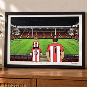 May include: A framed illustration of a family of three inside a football stadium. The figures wear red and white striped jerseys with the text MUM 10, DAD 1, and IVY 7. The text Our family. Our club. is printed at the bottom of the print.