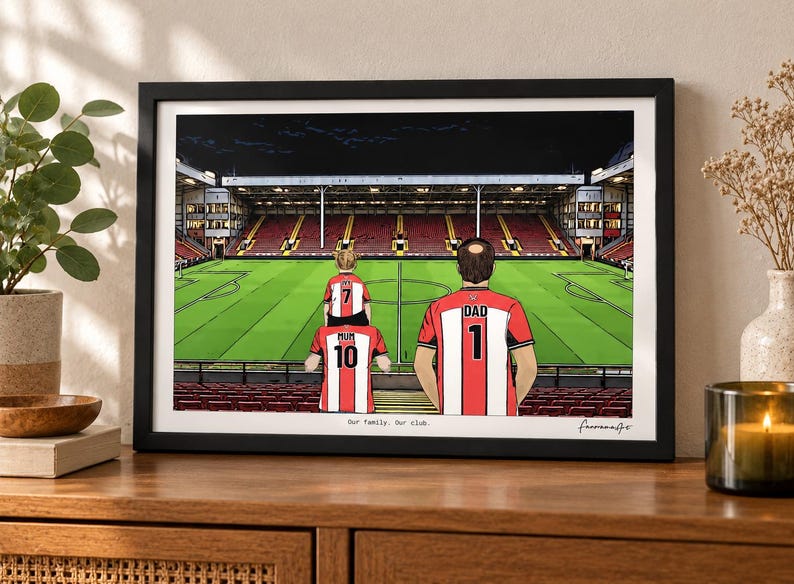 May include: A framed illustration of a family of three inside a football stadium. The figures wear red and white striped jerseys with the text MUM 10, DAD 1, and IVY 7. The text Our family. Our club. is printed at the bottom of the print.