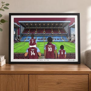 May include: A framed horizontal art print depicting a family of four wearing maroon and light blue soccer jerseys inside a stadium. The jerseys feature names and numbers: Dad 14, Max 7, Mum 10, and Ava 9. The text "Our Family. Our Club." is printed below.