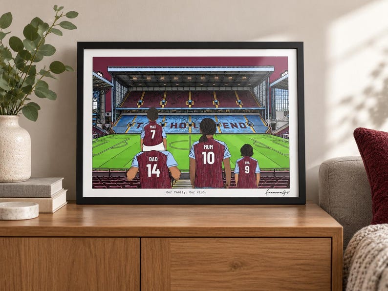 May include: A framed horizontal art print depicting a family of four wearing maroon and light blue soccer jerseys inside a stadium. The jerseys feature names and numbers: Dad 14, Max 7, Mum 10, and Ava 9. The text "Our Family. Our Club." is printed below.