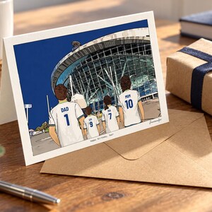 May include: A greeting card featuring a colorful illustration of four people wearing white soccer jerseys with blue trim, standing before a large stadium. The jerseys are labeled DAD 1, LEO 9, MIA 7, and MUM 10. The text Happy Birthday, Dad is printed below.