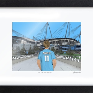 May include: A framed illustration of a stadium with a person in a light blue jersey with the name CHARLIE and the number 11 on the back. The text Our club. Our memories. is printed below the illustration in a black frame with a white mat.