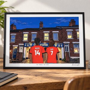 May include: A framed art print depicting two individuals wearing red soccer jerseys with the names Alex and Sam and numbers 14 and 7 on the back. They stand before a brick building with a blue sign reading Blocks Gate F G O Stand 7 D E.