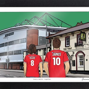 Pu&ograve; includere: Un'illustrazione digitale incorniciata mostra due figure viste di spalle che indossano maglie da calcio rosse con i nomi ELLIE e JAMES e i numeri 8 e 10 stampati sul retro. Lo sfondo presenta uno stadio e un edificio chiamato THE TURF.