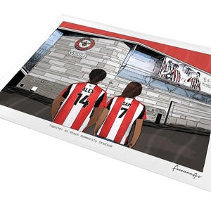 May include: A digital illustration print showing two individuals from behind wearing red and white striped football shirts with the names ALEX 14 and SAM 7, standing before the Gtech Community Stadium with the text Together at Gtech Community Stadium.