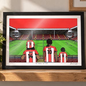 May include: A framed illustration of a family of four wearing red and white striped soccer jerseys with names and numbers on the back, standing in a stadium. The text "Our family. Our club." is printed at the bottom of the artwork.