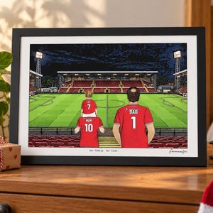 Pu&ograve; includere: Un'illustrazione incorniciata di uno stadio di calcio di notte con tre figure in maglie rosse con il testo "MUM 10", "DAD 1" e "IVY 7" sul retro. La stampa include il testo "Our Family. Our club." in basso al centro.