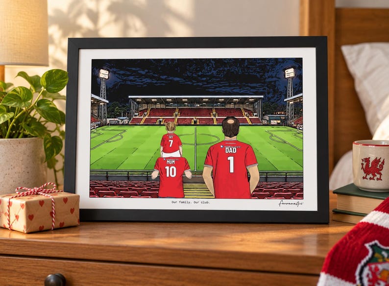 Pu&ograve; includere: Un'illustrazione incorniciata di uno stadio di calcio di notte con tre figure in maglie rosse con il testo "MUM 10", "DAD 1" e "IVY 7" sul retro. La stampa include il testo "Our Family. Our club." in basso al centro.