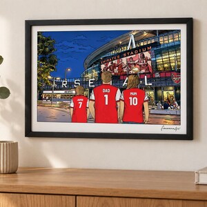 AI-created lifestyle mockup of an A4 Arsenal family print in a black frame on a cream wall above an oak sideboard, featuring Jamie 7, Dad 1 and Mum 10 at Emirates Stadium at night.