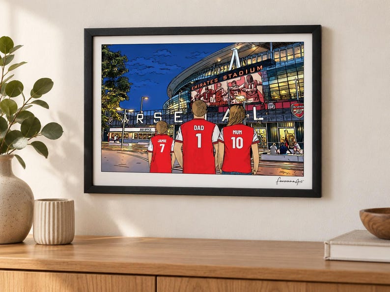 AI-created lifestyle mockup of an A4 Arsenal family print in a black frame on a cream wall above an oak sideboard, featuring Jamie 7, Dad 1 and Mum 10 at Emirates Stadium at night.