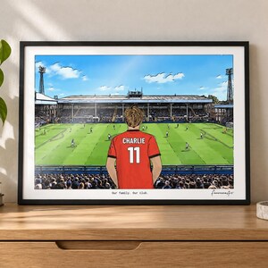 May include: A framed horizontal art print depicting a soccer stadium scene with a person wearing a red jersey with the name CHARLIE and the number 11 on the back. The text Our family. Our club. is printed at the bottom of the illustration.