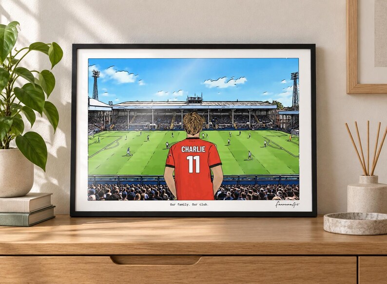 May include: A framed horizontal art print depicting a soccer stadium scene with a person wearing a red jersey with the name CHARLIE and the number 11 on the back. The text Our family. Our club. is printed at the bottom of the illustration.