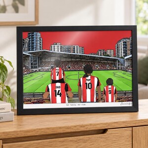 May include: A framed illustration of a family of four wearing red and white striped football shirts inside a stadium. The shirts feature the names Max, Dad, Mum, and Ava with numbers 7, 14, 10, and 9. The text "Our family. Our club." is printed below.