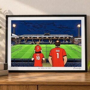 May include: A framed illustration of a soccer stadium at night featuring three figures in red jerseys with the text "MUM 10", "IVY 7", and "DAD 1" on their backs. The print includes the text "Our family. Our club." at the bottom center.