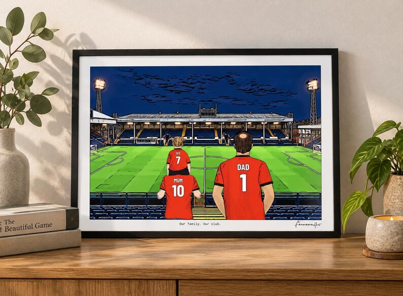 May include: A framed illustration of a soccer stadium at night featuring three figures in red jerseys with the text "MUM 10", "IVY 7", and "DAD 1" on their backs. The print includes the text "Our family. Our club." at the bottom center.
