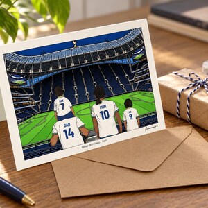May include: A greeting card featuring an illustration of four people in white soccer jerseys with blue numbers and names sitting in a stadium. The jerseys are labeled Max 7, Dad 14, Mum 10, and Joe 9. The text at the bottom reads Happy Birthday, Dad!