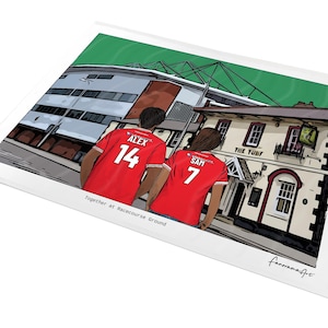Pu&ograve; includere: Una stampa con illustrazione digitale che mostra due persone che indossano maglie da calcio rosse con i nomi ALEX 14 e SAM 7 sul retro. Si trovano davanti a un edificio chiamato THE TURF e a uno stadio, con il testo Together at Racecourse Ground.
