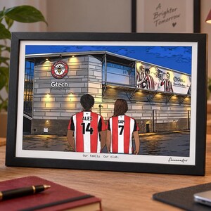 May include: A framed illustration of the Gtech Community Stadium featuring two people from behind wearing red and white striped football shirts with the names ALEX 14 and SAM 7. The text reads Gtech Community Stadium and Our family. Our club.