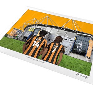 May include: A digital illustration print featuring two individuals wearing black and amber striped football shirts with the names ALEX 14 and SAM 7 on the back, standing before a stadium with an orange sky. The text reads Together at MKM Stadium.