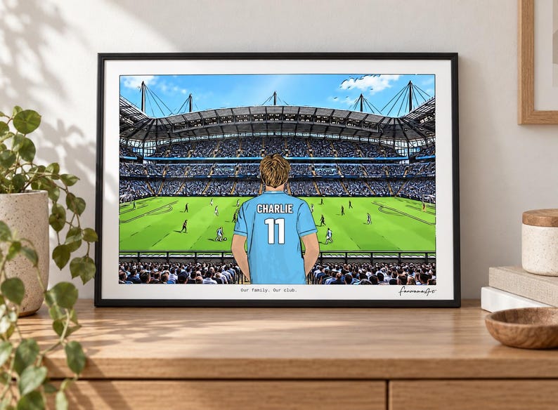 May include: A framed illustration of a soccer stadium featuring a person wearing a light blue jersey with the name CHARLIE and the number 11 on the back. The text Our family. Our club. is printed at the bottom center of the artwork.