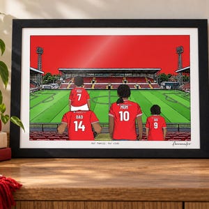 Pu&ograve; includere: Un'illustrazione incorniciata di una famiglia di quattro persone che indossa maglie da calcio rosse con nomi e numeri in uno stadio. Le maglie riportano le scritte Dad 14, Max 7, Mum 10 e Ana 9. Il testo Our family. Our club. &egrave; stampato in basso.