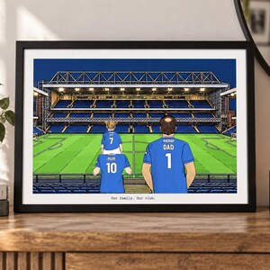 May include: A framed illustration of a soccer stadium with three people in blue jerseys standing in the foreground. The jerseys are labeled Mum 10, Dad 1, and a child with 7. The text Our family. Our club. is printed at the bottom of the print.