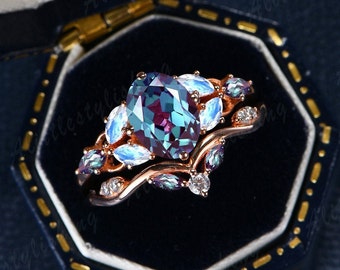 May include: A rose gold engagement ring set with a large oval alexandrite center stone surrounded by smaller alexandrite and moonstone accent stones. The ring is in a blue velvet ring box.