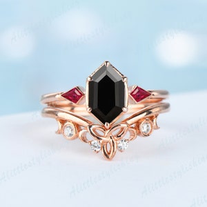 Könnte beinhalten: Ein Verlobungsring aus Roségold mit einem schwarzen, achteckigen Edelstein, zwei kleinen roten Edelsteinen und einem filigranen Schmetterlingsdesign mit kleinen weißen Edelsteinen.