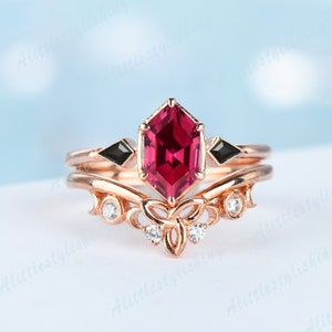 May include: A rose gold ring set featuring a hexagonal, faceted, ruby-coloured gemstone as the centerpiece. The engagement ring has black diamond accents and the wedding band has a floral design with small, clear gemstones.